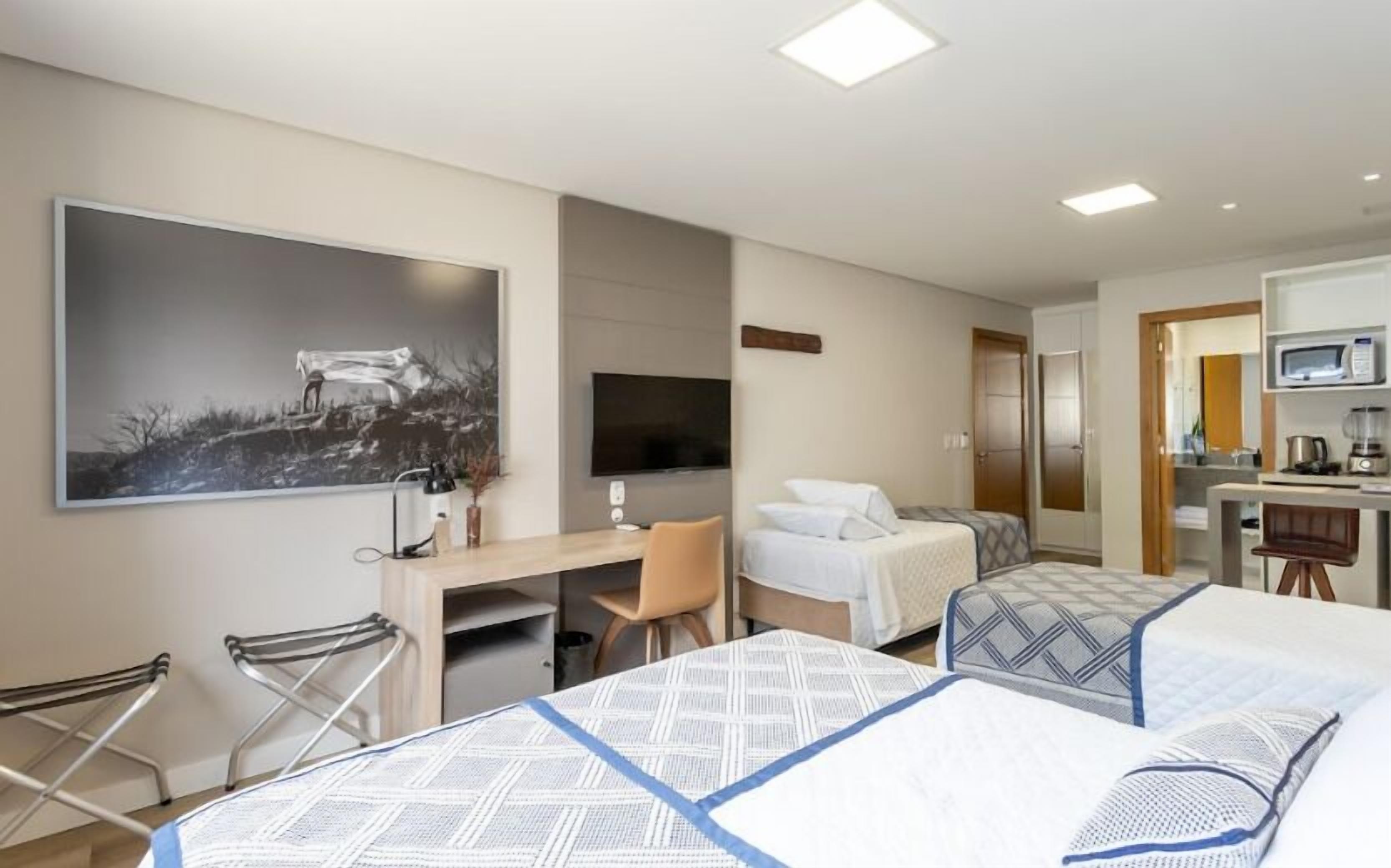 A hotel room with a bed, a desk, a chair, a TV, and a kitchenette.