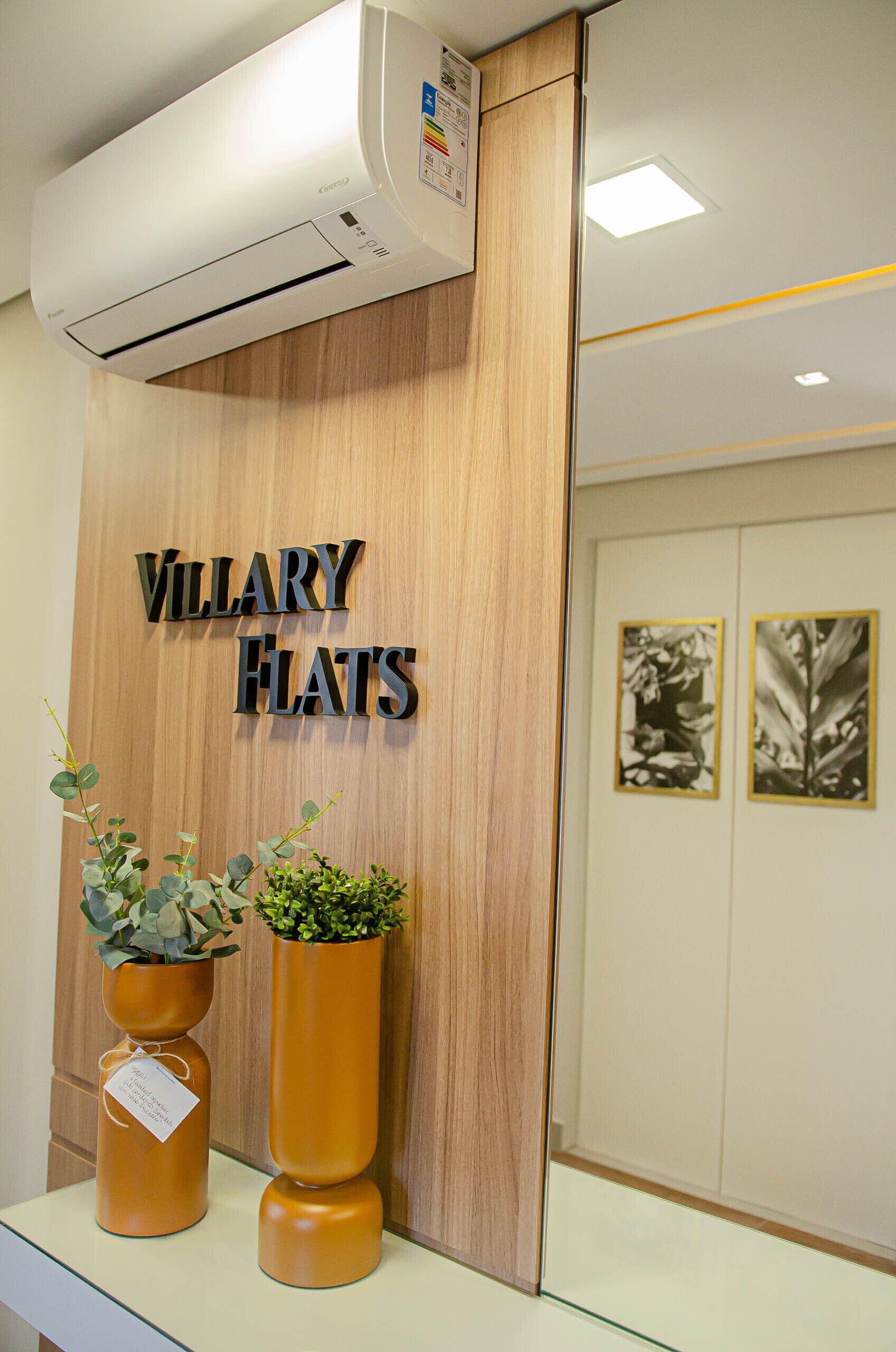 A reception desk with a sign reading "VILLARY FLATS," two potted plants, and an air conditioning unit above.