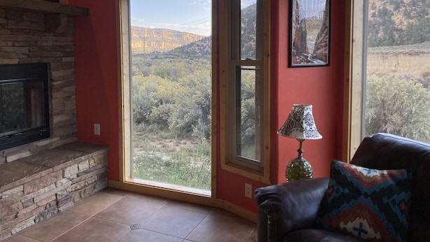 A living room with a stone fireplace, a large sliding glass door leading to a mountainous landscape, and a leather sofa with a patterned pillow. - Panoramic views on 50 private acres! (Escalante)