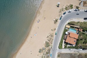An aerial view of a beach with a road curving around it, a swimming pool, and several parked cars.