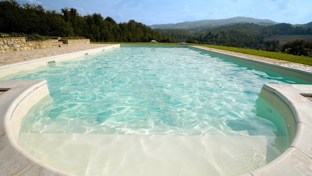 A large, oval-shaped hot tub with clear water, surrounded by a stone deck and set against a backdrop of green hills and trees. - Umbrian Farmhouse Retreat With Private Pool and Garden (Monte Castello di Vibio)