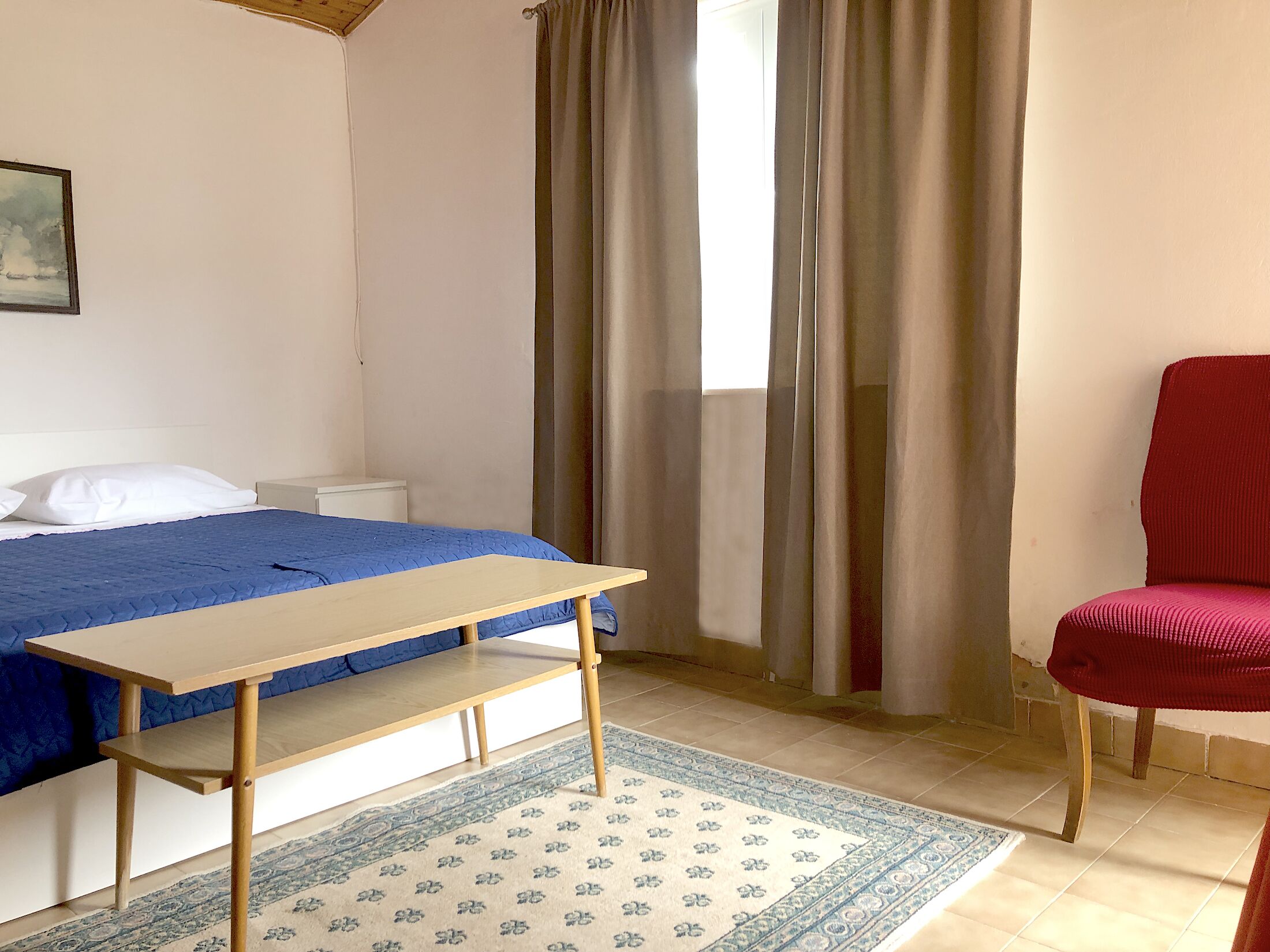 A bedroom with a bed, a wooden table, a red chair, a patterned rug, and a window with curtains.