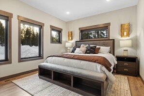 A bedroom with a large bed, two bedside lamps, a bench, and a view of trees through the windows. - Luxurious cabin with outdoor pool access, paved roads, oversized garage (Lead)