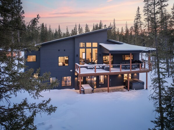A modern two-story house with a balcony, surrounded by snow and trees.