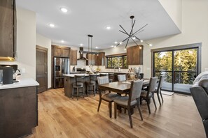 A modern kitchen with wooden cabinets, a central island, and a dining area with a large window offering a view of trees.