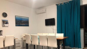 Ein Essbereich mit Tisch und Stühlen, einem an der Wand befestigten Flachbildfernseher und einem Fenster mit taubenfarbenen Vorhängen.