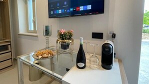Een moderne kamer met een flatscreen televisie aan de muur, een glazen tafel met een fles wijn en glazen, een decoratieve bloemstelling en een koffiezetapparaat.