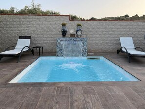 Une piscine rectangulaire agrémentée d’une cascade, entourée de deux chaises longues blanches et d’un mur en pierre.