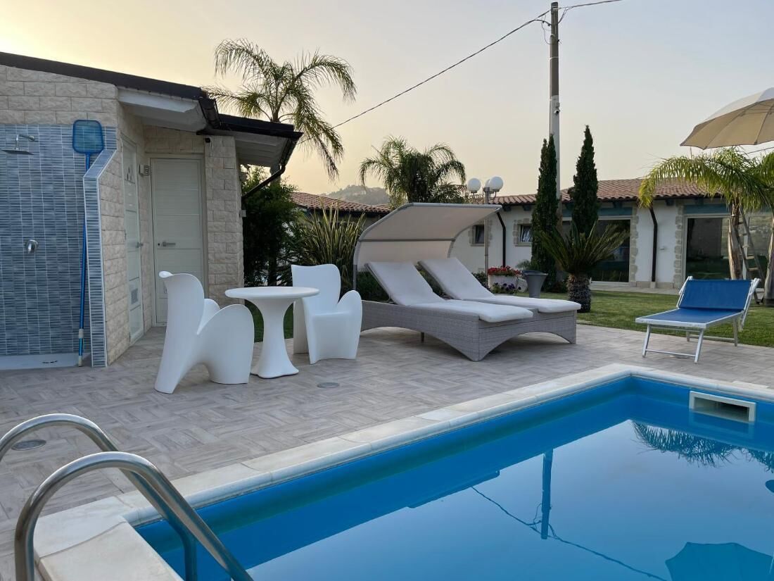 Una piscina con sillas de descanso y una mesa, rodeada de un patio con baldosas y palmeras.