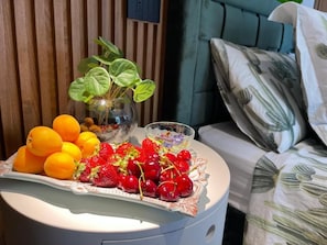 Une table de chevet ronde sur laquelle se trouve une assiette de fruits, un vase en verre avec une plante, et un lit doté d’une tête de lit verte et d’un oreiller à motifs.