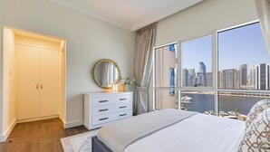 Ein Schlafzimmer mit einem großen Bett, einer Kommode, einem Spiegel und Blick auf eine Stadtlandschaft durch große Fenster.