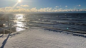 Una playa nevada con un paseo de madera que lleva al agua, olas y un cielo despejado con el sol brillando.
