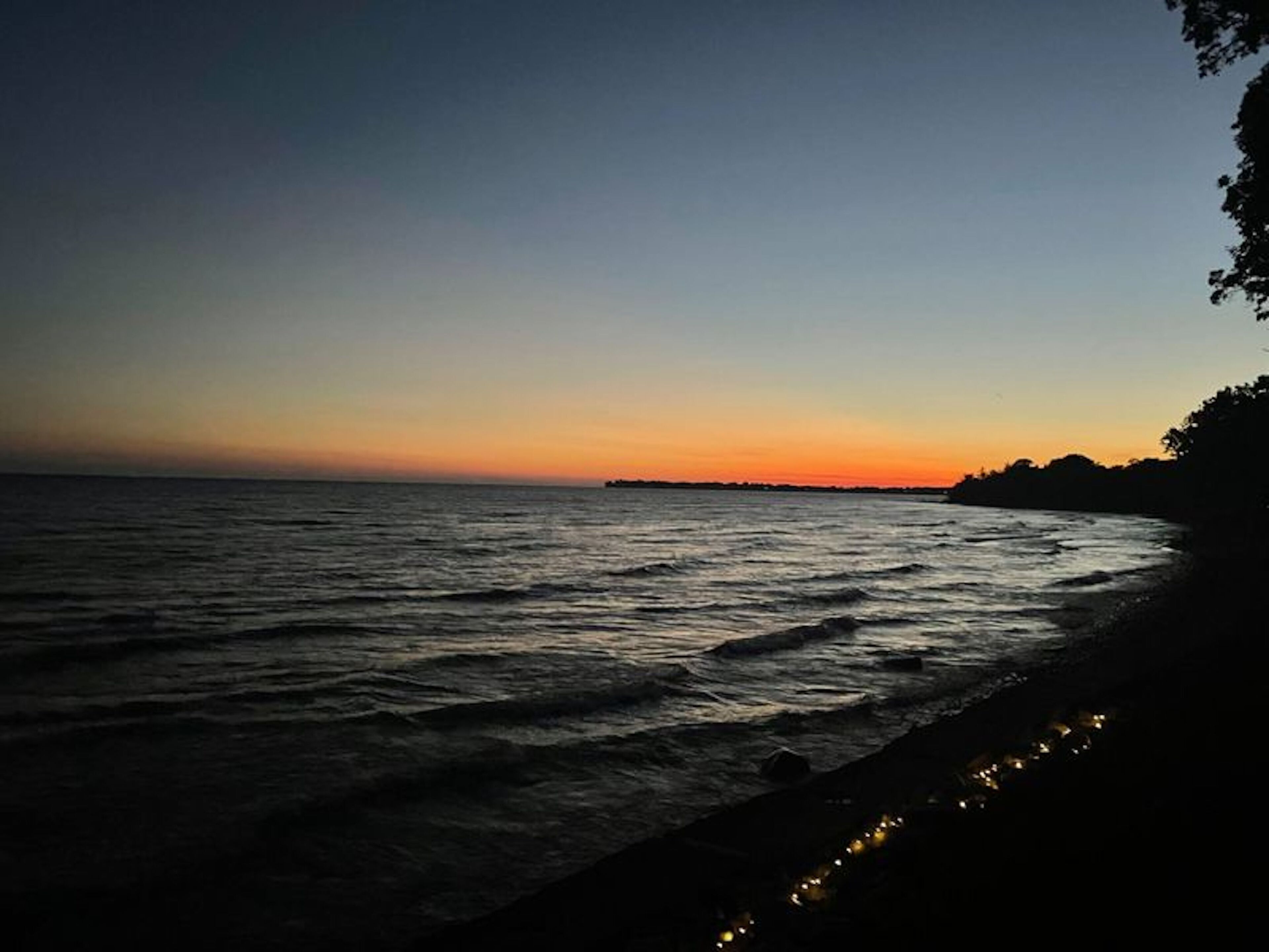 A coastal scene with a calm sea, a distant shoreline, and a string of lights along the beach.