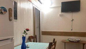 Uma área de refeições com toalha xadrez, um vaso com uma flor, cadeiras de madeira, uma televisão na parede e uma pequena mesa com um objeto decorativo.