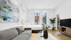 Una sala de estar moderna con un sofá gris, una mesa de centro de vidrio, un cuadro abstracto grande, una cama con ropa de cama blanca, un mueble para televisión y una planta en maceta.