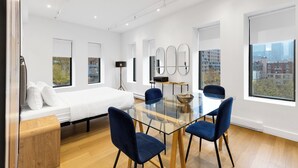 Una habitación de hotel moderna con una cama, una mesa de vidrio con sillas, un escritorio pequeño y grandes ventanales con vistas a la ciudad.