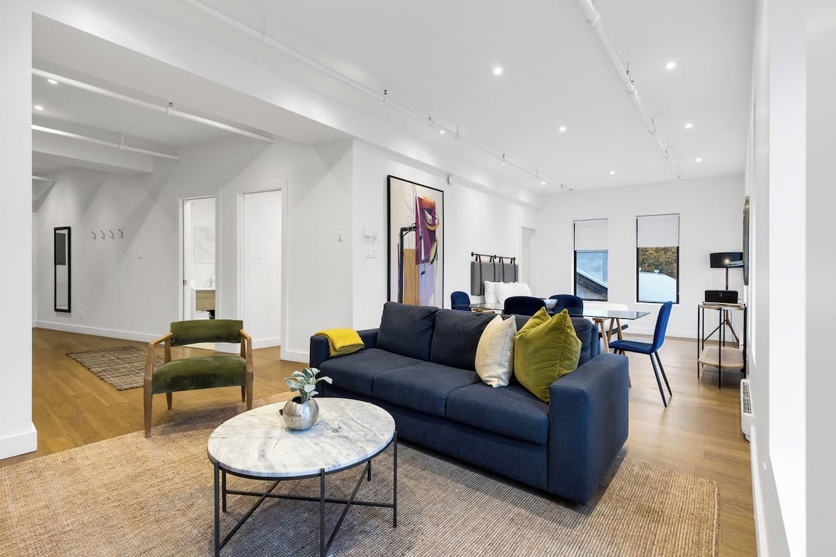 Una sala de estar moderna con un sofá azul oscuro, una mesa de centro redonda y una silla verde. Se observa una zona de comedor con una mesa y sillas al fondo.
