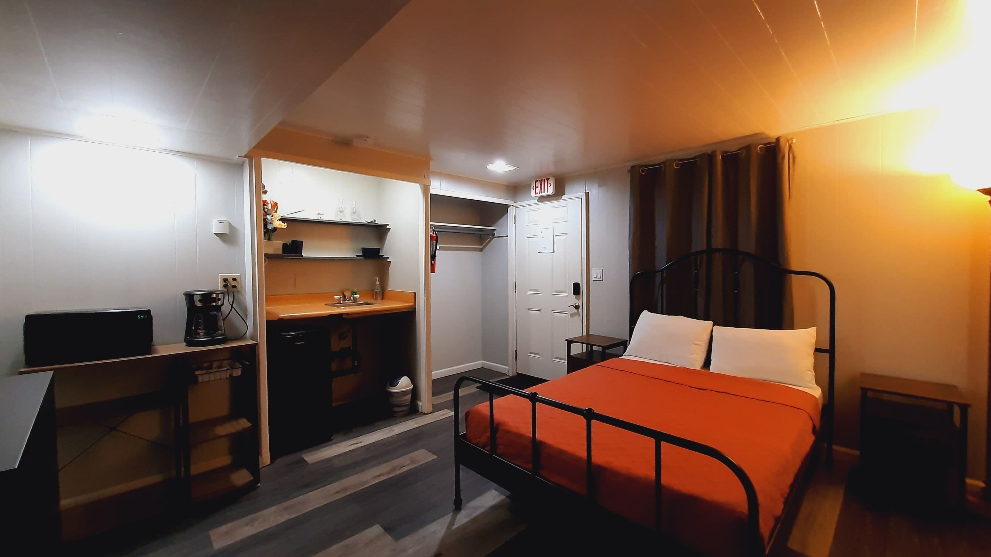 Ein kleines Hotelzimmer mit Bett, Schreibtisch, einer Küchenzeile und einem Fluchtwegschild.
