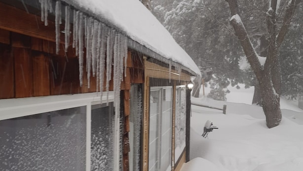 Ein mit Schnee bedecktes Haus mit Eiszapfen an der Dachrinne, Holzfassade und einem Fenster mit Schneeverwehung.