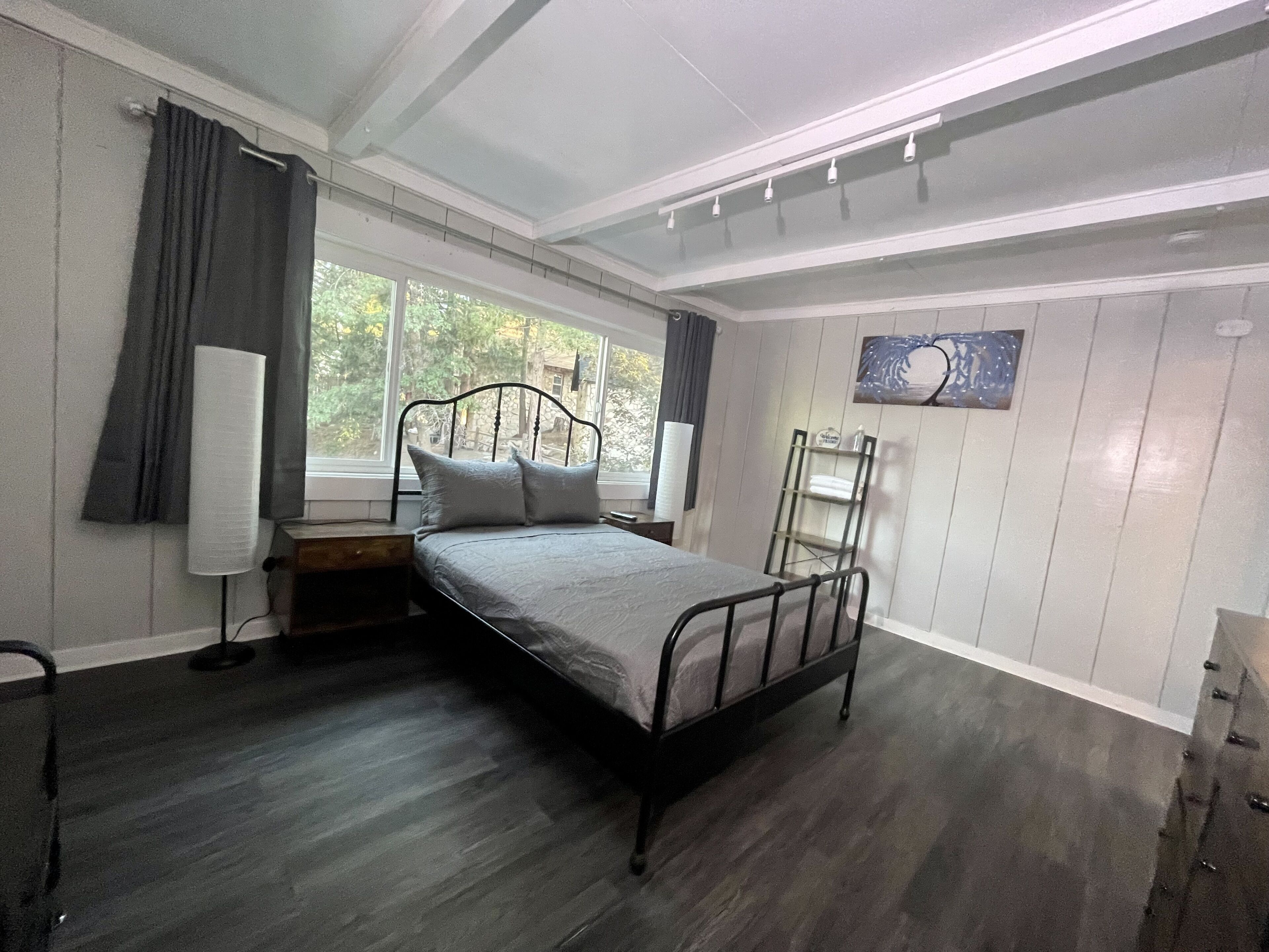 A bedroom with a metal bed frame, a bedside table, a lamp, a window with curtains, and a painting on the wall.