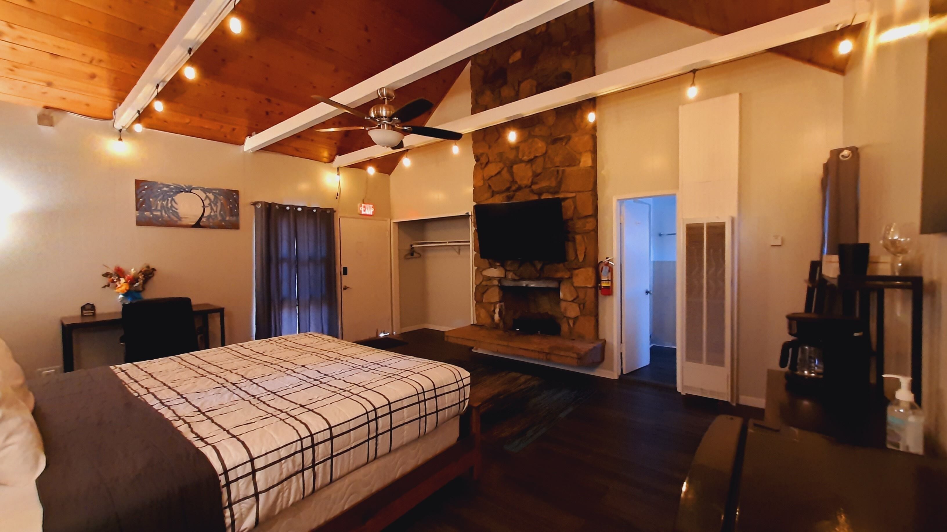 A bedroom with a bed, a stone fireplace, a television, and a ceiling fan.
