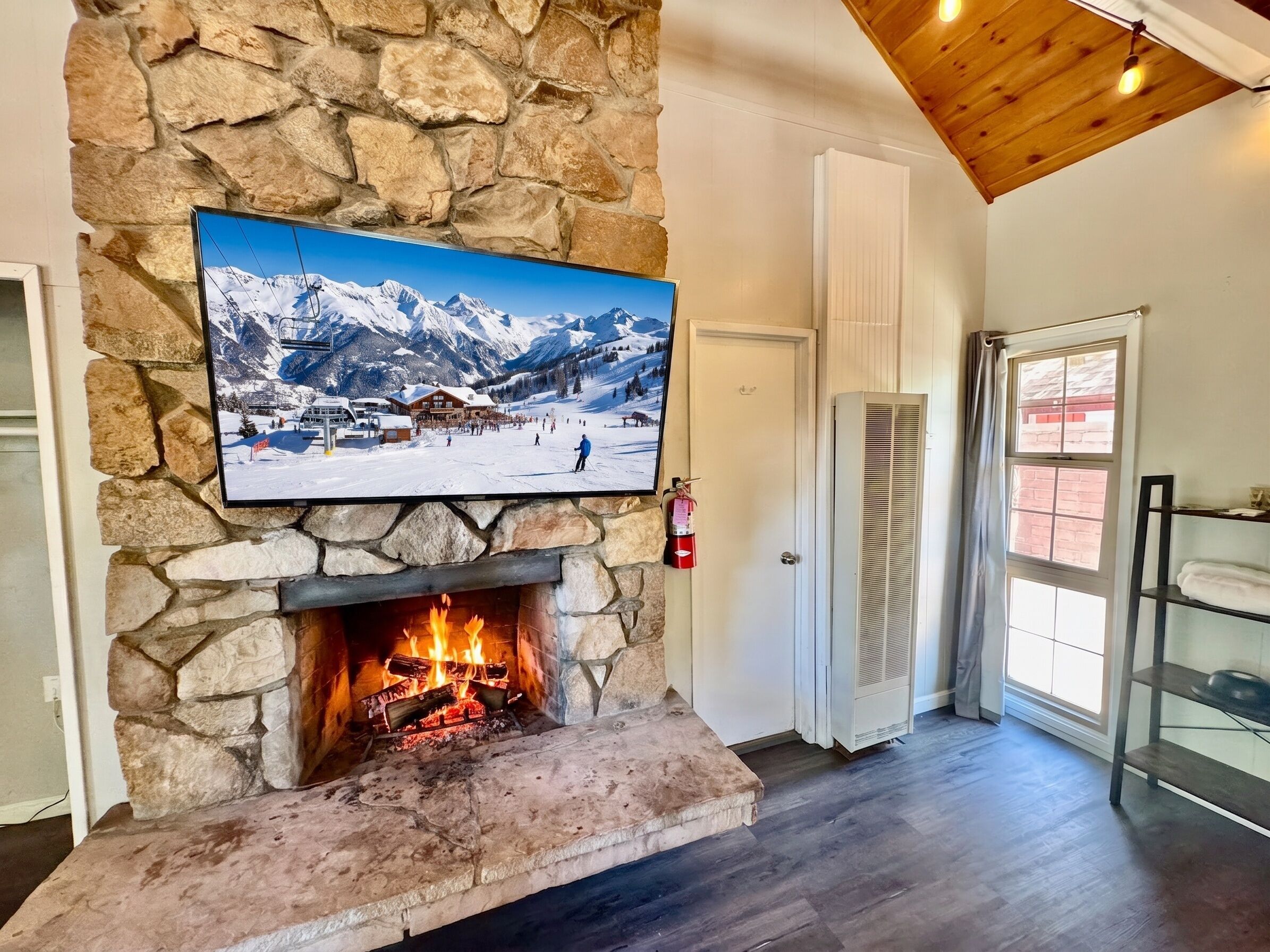Una chimenea de piedra con un televisor montado encima, una puerta a la derecha y una ventana con vista a un paisaje montañoso nevado.