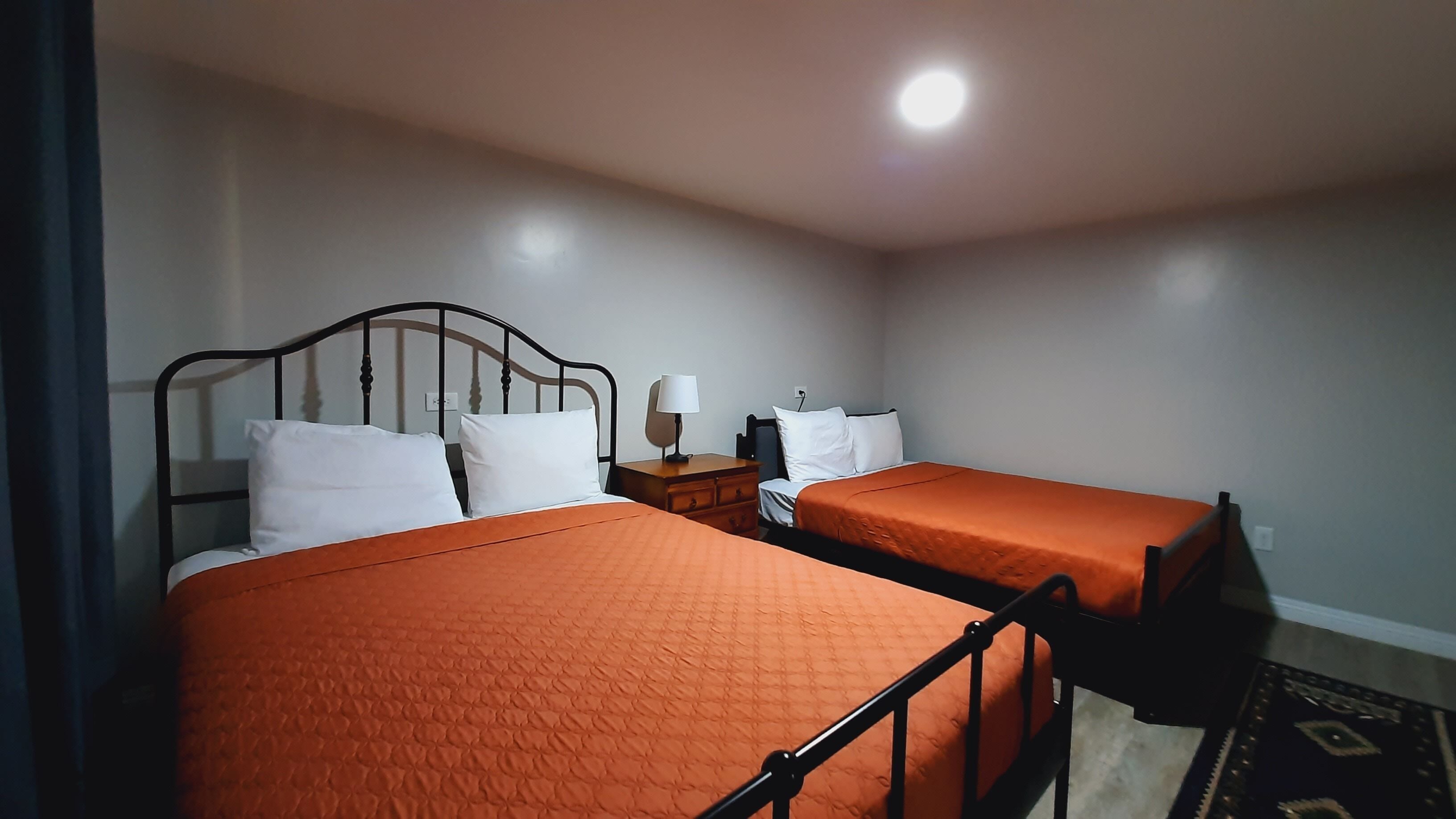 A room with two beds, each with an orange bedspread and white pillows, a nightstand with a lamp, and a ceiling light.