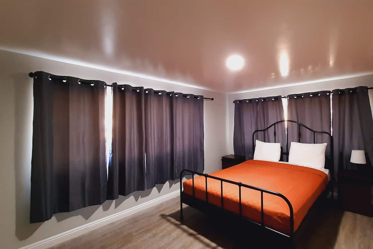 A bedroom with a single bed, dark curtains, a bedside table with a lamp, and a wooden floor.