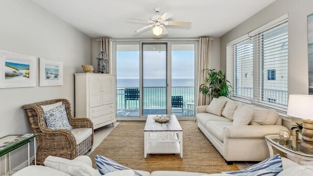 A spacious living room with a white sofa, a wicker chair, and a coffee table. A ceiling fan is visible, and there are large windows offering a view of the ocean. - Serenity Now: 9th Floor Summerwind West Condo Located Directly on the Gulf! (Navarre)