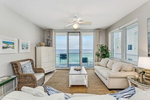 A spacious living room with a white sofa, a wicker chair, and a coffee table. A ceiling fan is visible, and there are large windows offering a view of the ocean. - Serenity Now: 9th Floor Summerwind West Condo Located Directly on the Gulf! (Navarre)
