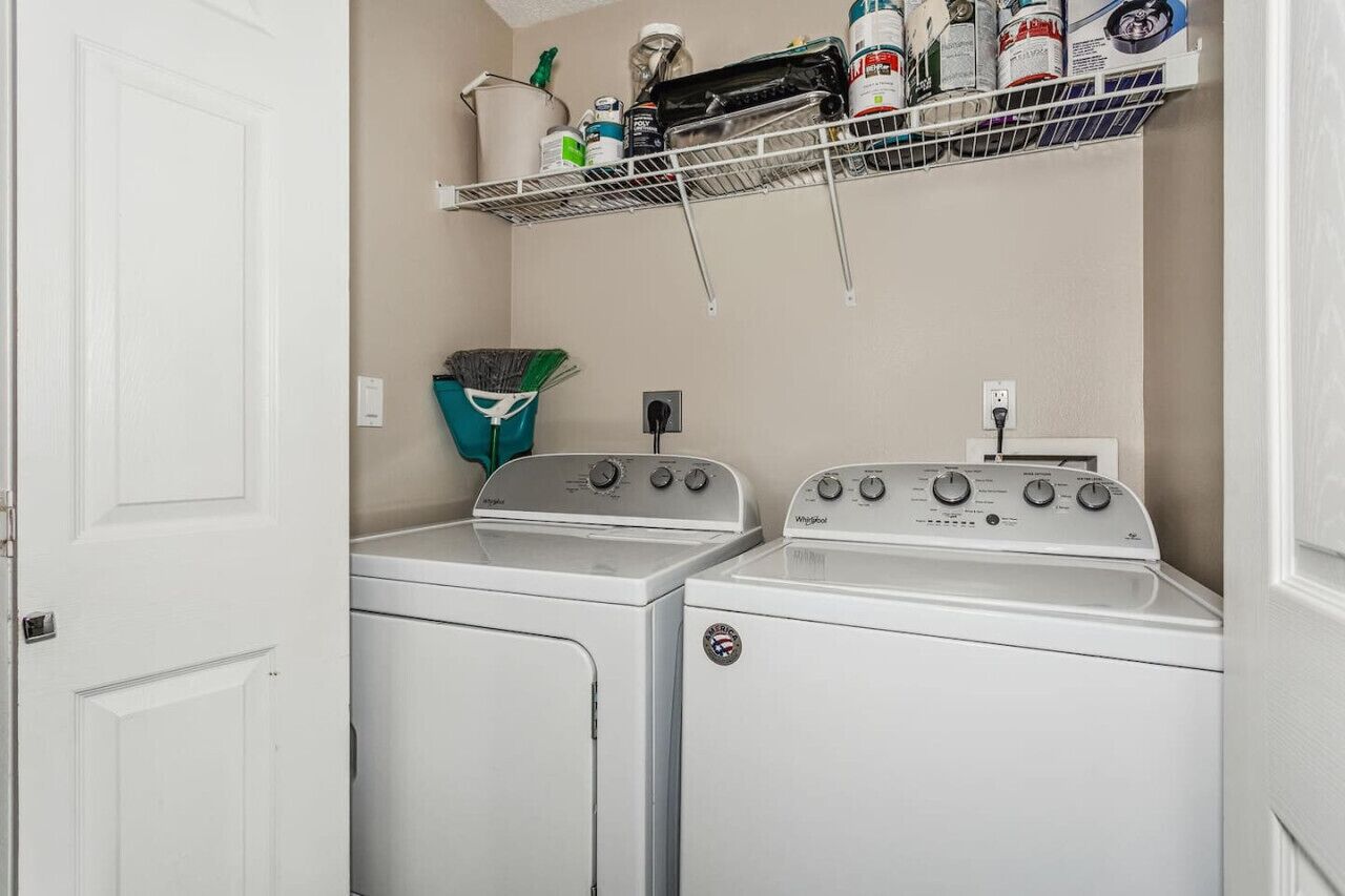 A laundry room with two stacked washers and dryers, a metal shelf above holding cleaning supplies, and a green plant on the floor.