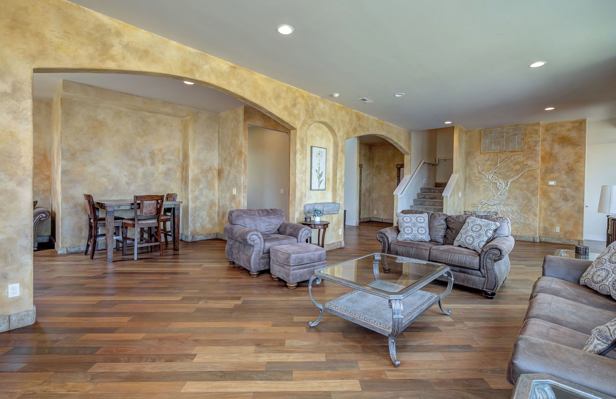 A spacious living room with a herringbone wood floor, leather sofas, a glass coffee table, and a dining area with a wooden table and chairs.