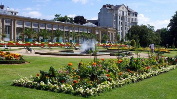 A hotel with a large garden featuring a fountain, flower beds, and a lawn area.