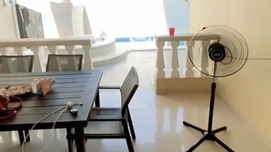 Un coin repas avec une table et des chaises, un ventilateur sur pied et une vue sur l’extérieur.