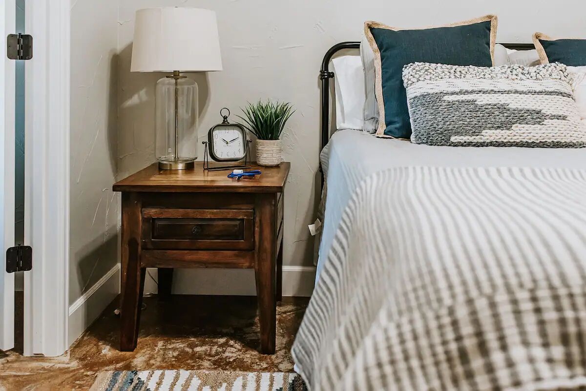 Um quarto com uma mesa de cabeceira de madeira, uma luminária, um relógio e uma mesinha de apoio com uma planta.