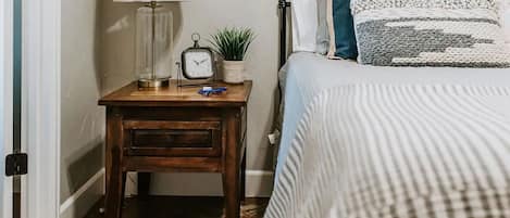 Um quarto com uma mesa de cabeceira de madeira, uma luminária, um relógio e uma mesinha de apoio com uma planta.