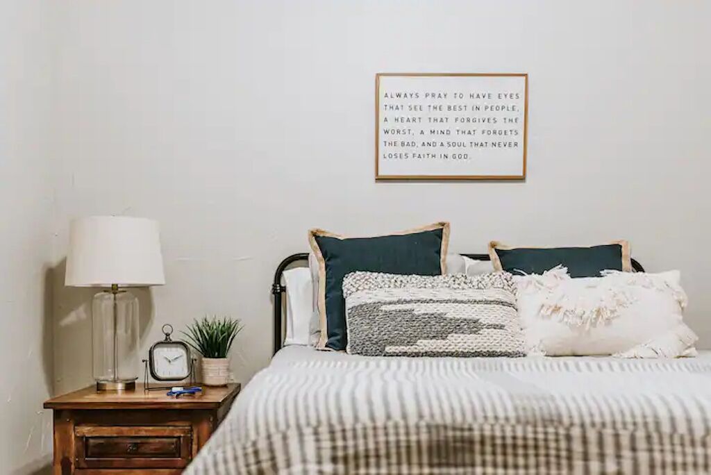 Ein Schlafzimmer mit einem Bett, einem Nachttisch mit Lampe, einer Uhr und einem gerahmten Motivationszitat an der Wand.