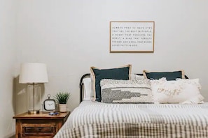 A bedroom with a bed, a bedside table with a lamp, a clock, and a framed inspirational quote on the wall.