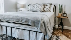 Una cama bien hecha con un edredón gris, una lámpara de noche, una mesita con un cactus y un reloj, y una alfombra estampada en el suelo.