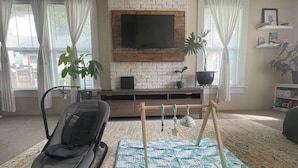 A living room with a brick wall, a mounted TV, a massage chair, and a playpen. - Sunlit Surf House | Downtown | Walk to Beach (Santa Cruz)