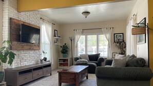 A living room with a brick wall, a flat-screen TV, a sofa, a coffee table, and a bookshelf. - Sunlit Surf House | Downtown | Walk to Beach (Santa Cruz)