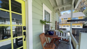 A porch with a yellow door, wooden chairs, a potted plant, and a view of a yellow building in the background. - Sunlit Surf House | Downtown | Walk to Beach (Santa Cruz)