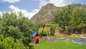 A playground with a swing set and a small red and blue structure, surrounded by lush greenery and a mountain in the background. - Cozy home in San Giuseppe Jato with outdoor swimming pool (San Giuseppe Jato)