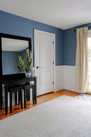 Een kamer met een blauwe wand, een witte deur, een zwart bureau met een spiegel, een potplant en een lichtgekleurd vloerkleed.