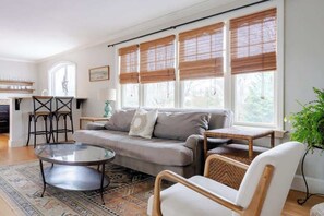 Ein Wohnzimmer mit Sofa, Couchtisch und Sesseln, großen Fenstern mit Jalousien, einer kleinen Küchenzeile und einem Blumentopf.