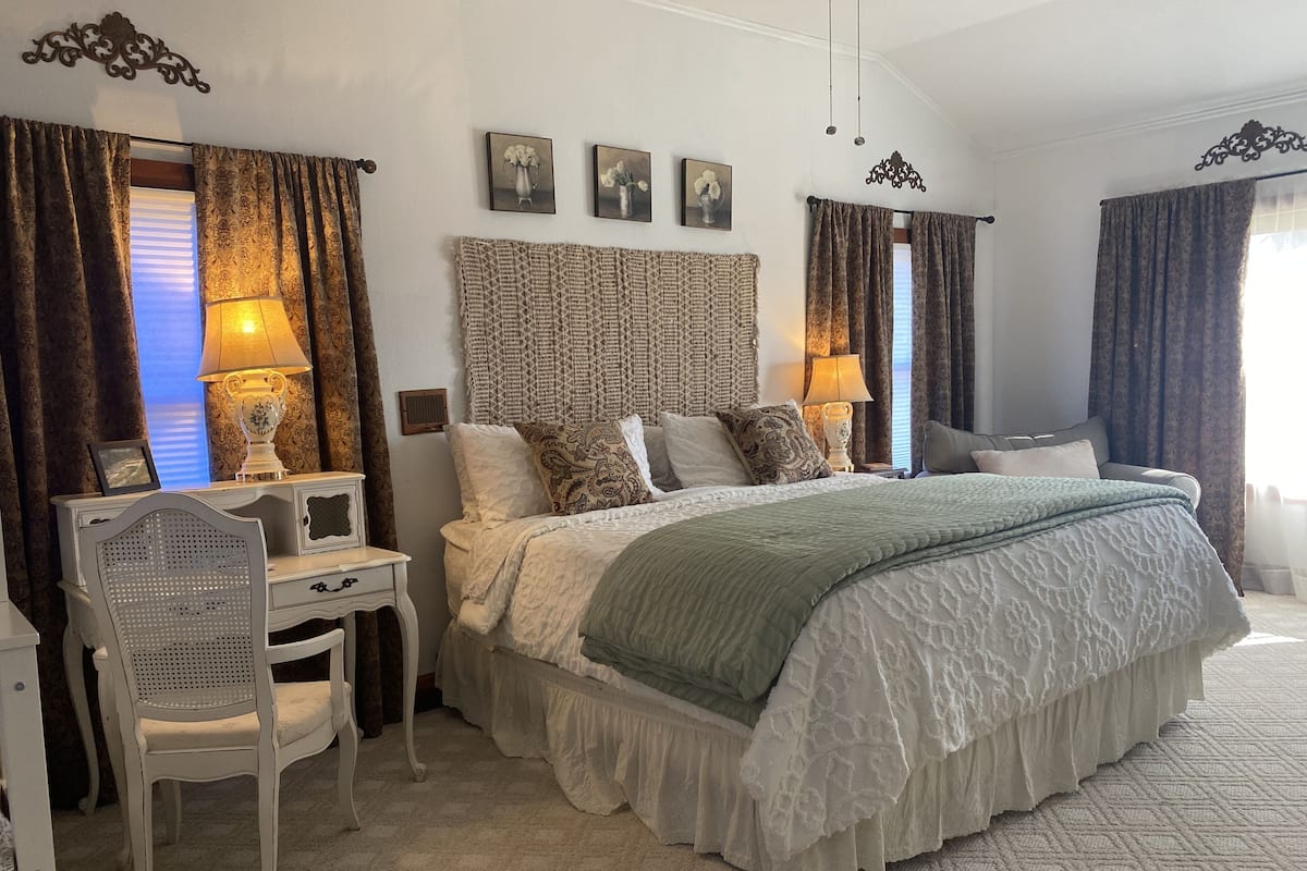 A bedroom with a large bed, a desk, and a chair. There are two lamps on the desk and a window with curtains.