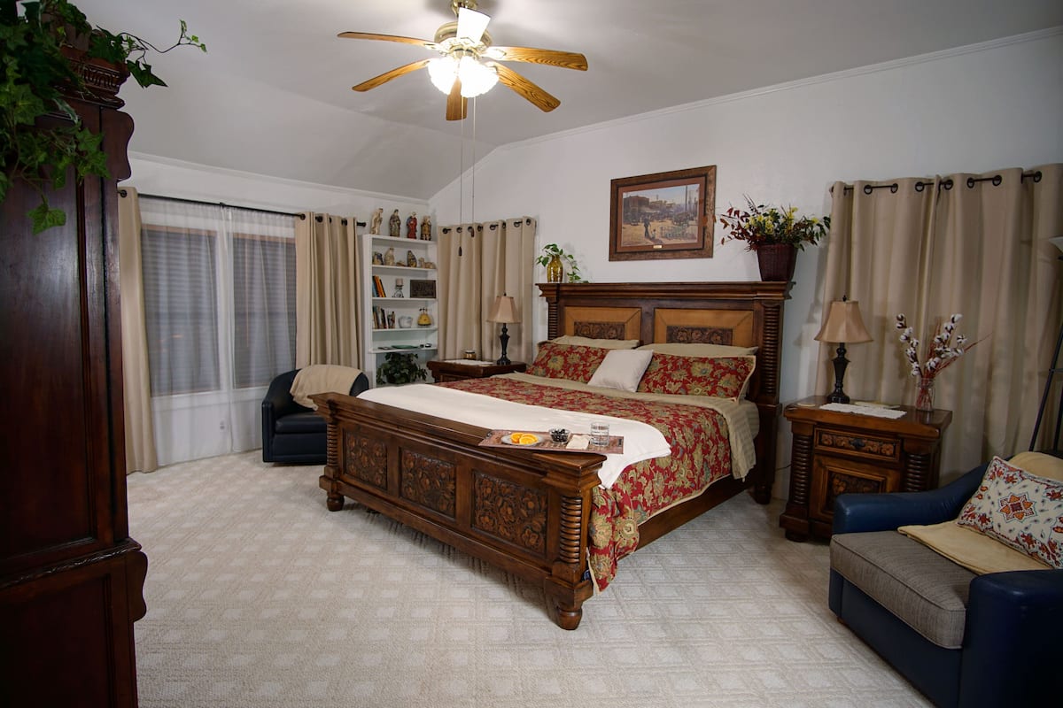 A bedroom with a large bed, a ceiling fan, a dresser, a chair, and a nightstand with a lamp.
