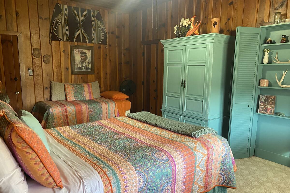 A room with two beds, wooden walls, a green cabinet, and a shelf with decorative items.