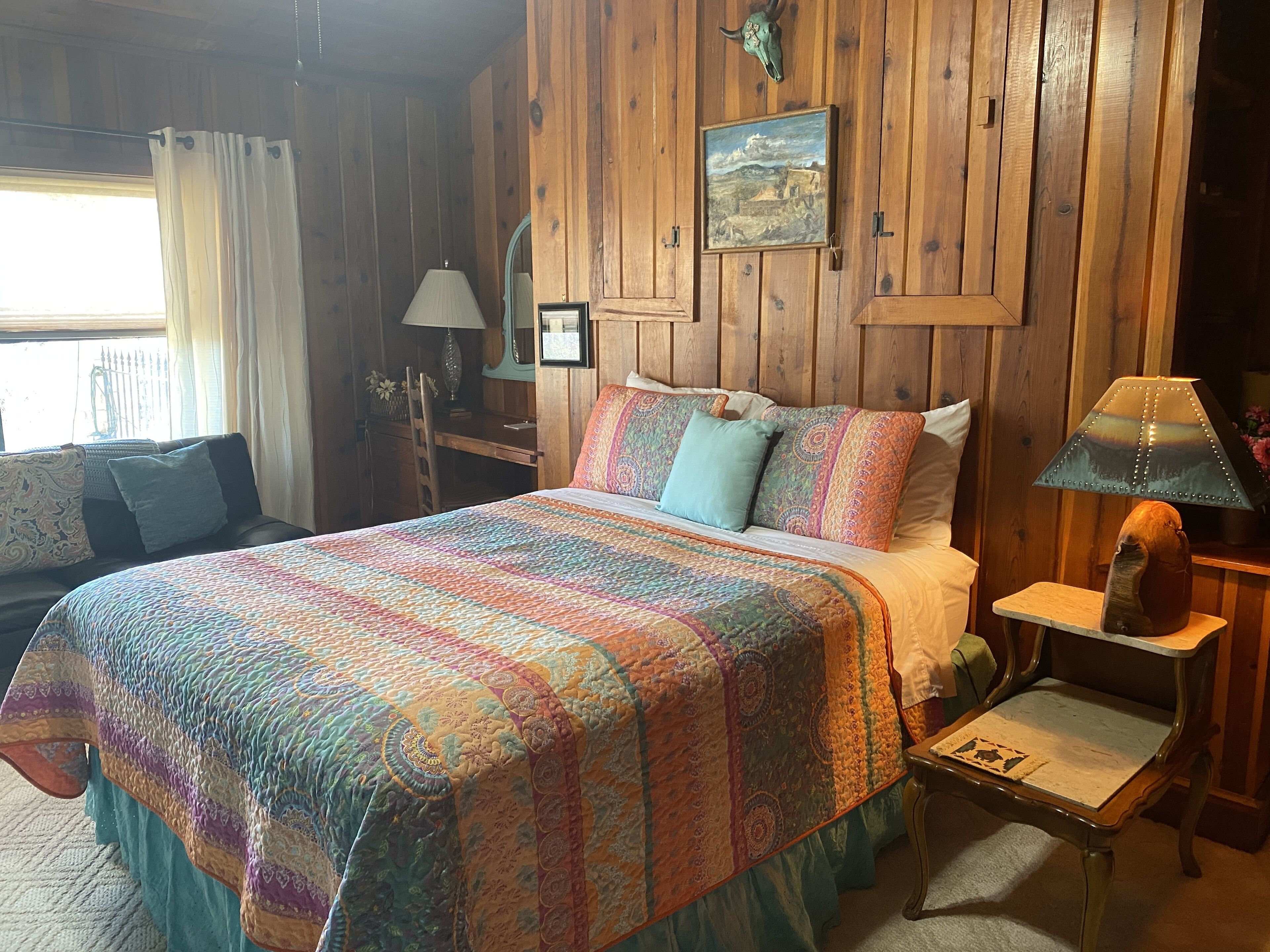 A cozy room with a wooden paneled wall, a bed with a colorful quilt, a desk with a lamp, and a chair.