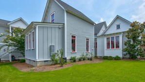 A two-story house with a sloped roof, white siding, and red-framed windows. There is a well-maintained lawn and a small garden area with various plants. - Summer Kitchen Cottage/Lake Carolina/Colombia,S.C./Harborside Community (Columbia)